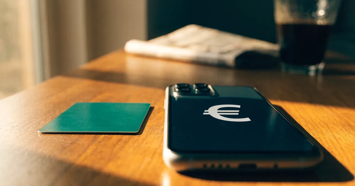 Um telemóvel e um cartão bancário lado a lado numa mesa de madeira, com algumas notas de euro ao lado